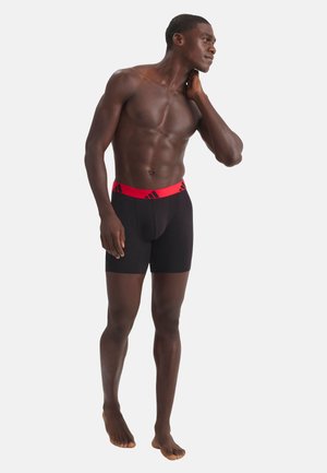 Hombre atlético de pie, descalzo y sin camiseta, vistiendo calzoncillos tipo bóxer negros con una banda roja en la cintura, mirando hacia un lado con una mano en el cuello.