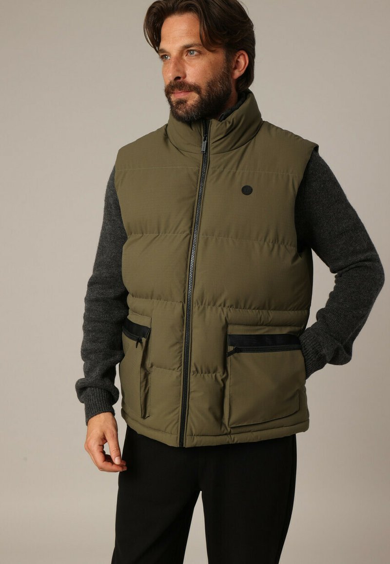 Olijfgroene gewatteerde vest met hoge kraag, zwarte accenten en twee vooraan aangebrachte ritszakken, gedragen over een grijze trui. Gladde textuur en gestructureerd ontwerp.