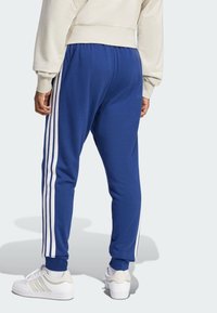 Pantalones de chándal azules con franjas laterales blancas, cinturilla elástica y un corte relajado, combinados con zapatillas blancas y un suéter de color beige claro.