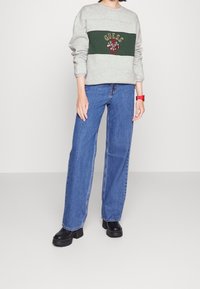 Světle šedá mikina s zeleným horizontálním pruhem, na kterém je vyšitý logo "GUESS", doplněná širokými modrými džínami a černými platformovými botami.