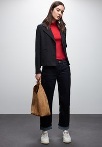 Giacca nera aderente sopra un dolcevita rosso, abbinata a jeans scuri e sneakers bianche. Tiene una borsa in camoscio beige. Design semplice e moderno.
