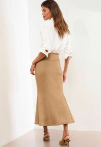 Jupe mi-longue beige en tissu doux, avec une taille ajustée et un bas évasé. Associée à une chemise blanche à boutons.