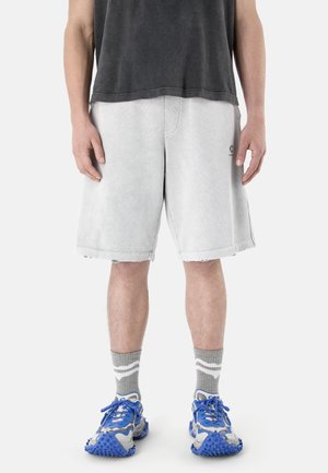 Mann trägt hellgraue knielange Shorts, graue Socken mit weißen Streifen, blaue und grau strukturierte Sneakers sowie ein dunkelgraues Hemd.