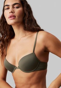 Oliwkowy stanik z lekko wyściełanymi miseczkami, regulowanymi ramiączkami i gładką fakturą. Posiada przeszycia oraz mały metalowy akcent.