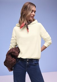 Vrouw draagt een crèmekleurige hoodie en een roodgepatroonde nekdoek terwijl ze een bruin doorgestikt jack vasthoudt, poseert tegen een lichtblauwe achtergrond.