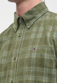 Grønn rutete skjorte laget av teksturert stoff, med krage med knapper, to knapper på placket og en liten logo i rødt, hvitt og blått.