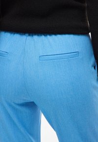 Heledad sinised püksid tekstuuriga kangast, millel on elastne vöökoht ja kaks tagumist taskut. Vaade on tagant.