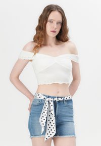 Top court blanc à épaules dénudées avec une bordure festonnée, associé à un short en denim bleu avec une ceinture à pois noir et blanc.