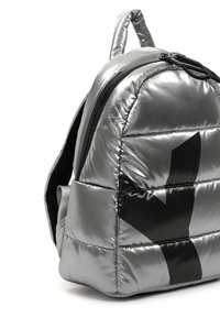 Sac à dos matelassé argenté brillant avec fermeture éclair noire, design abstrait noir et poignée rembourrée sur fond blanc.