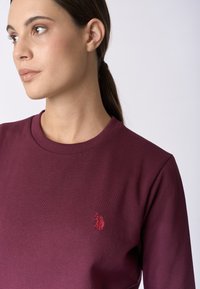 Felpa bordeaux con collo a costine e un piccolo logo rosso ricamato sul petto. Tessuto morbido, vestibilità comoda.