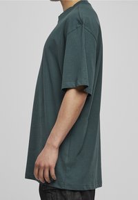 Vista laterale di una persona che indossa una maglietta oversized verde scuro a maniche corte e pantaloni scuri su uno sfondo grigio uniforme.