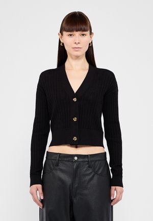 Schwarze kurz geschnittene Strickjacke mit gerippter Textur, V-Ausschnitt und vier großen goldfarbenen Knöpfen. Über schwarzen Lederhosen getragen.