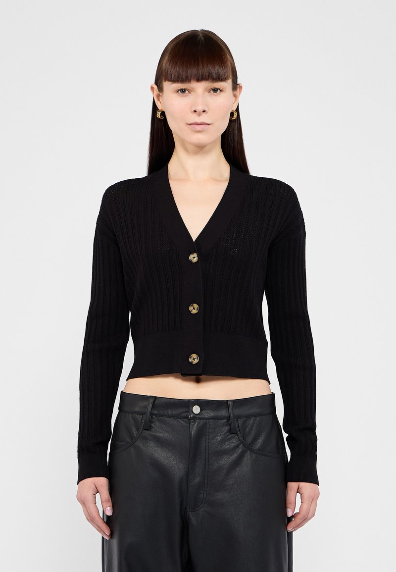 Cardigan noir court avec texture côtelée, encolure en V et quatre grands boutons en ton doré. Porté par-dessus un pantalon en cuir noir.