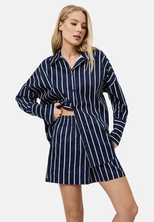 Vrouw draagt een marineblauw-wit verticaal gestreept overhemd met knopen en bijpassende short met elastische taille, staand met één hand in de zak.