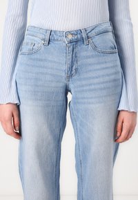 Világoskék denim farmernadrág magas derékkal, egyenes szárú fazonnal és minimális kopással. Kidolgozott varrott zsebekkel és fémből készült gombos zárással.
