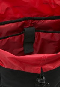 Bolsa negra con forro interior rojo, que cuenta con una correa central, cierre de cordón y una textura suave y duradera.