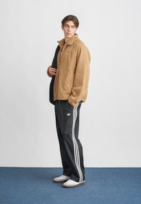 Fiatal személy bézs gyapjú dzsekit, fekete Adidas sportnadrágot fehér csíkokkal, fehér sportcipőt visel, és egy fekete táskát hordoz a válla fölött.