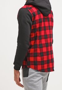 Camicia di flanella a scacchi rossi e neri con maniche lunghe nere, dotata di cappuccio. Il tessuto sembra morbido con una vestibilità comoda. Sono visibili pantaloni grigi.