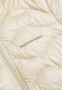 Beige quiltet jakke med en blød, skinnende tekstur, forsynet med lynlås i lodret retning og "Peak Performance"-logo i guldtråd.