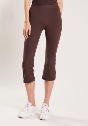 Donna che indossa leggings marroni corti e scarpe da ginnastica bianche, in piedi contro uno sfondo chiaro e semplice.