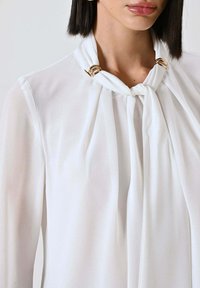 Camicia bianca realizzata in tessuto leggero e trasparente con colletto legato con dettagli ad anello dorato. Maniche lunghe e vestibilità morbida.