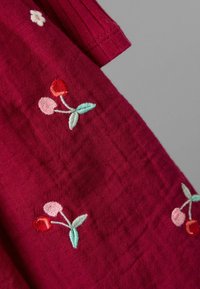 Burgunderfarbener Stoff mit gestepptem Muster, das florale Stickereien in Rot-, Rosa- und Grüntönen aufweist. Kurzer, gerippter Kragen ist sichtbar.