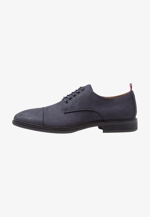 Chaussure Oxford en daim bleu marine à lacets, bout droit, semelle noire et languette rouge au talon, vue de profil.