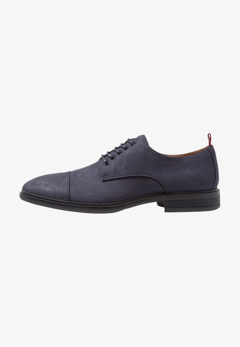Chaussure Oxford en daim bleu marine à lacets, bout droit, semelle noire et languette rouge au talon, vue de profil.