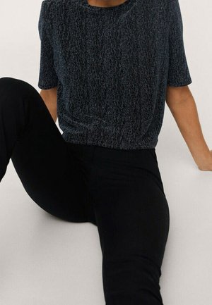 Osoba siedząca z prawą nogą zgiętą, a lewą wyciągniętą, ubrana w ciemną, teksturowaną koszulkę z krótkim rękawem i czarne spodnie na jednolitym tle.