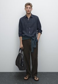 Man wearing a dark blue shirt, tied sweater at waist, brown pants, and flip-flops, holding a large dark bag, standing against a plain wall.