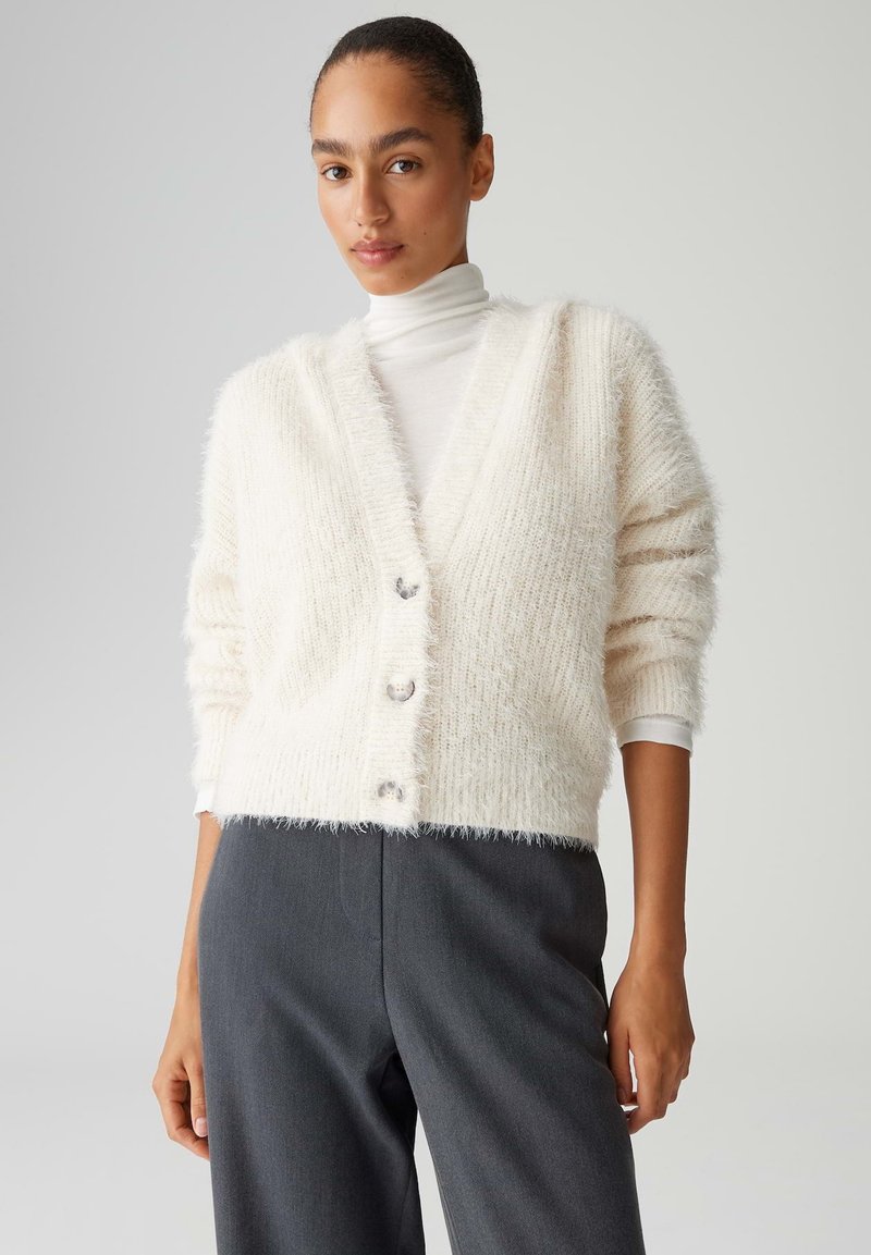 Weißer flauschiger Cardigan mit V-Ausschnitt, Knopfleiste und dreiviertellangen Ärmeln, kombiniert mit grauen, hoch taillierten Hosen. Weiche Textur.