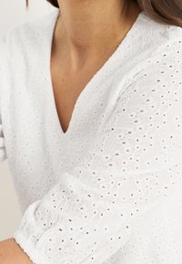 Biała bluzka z haftowanymi otworami, dekoltem w kształcie litery V i marszczonymi mankietami. Lekka tkanina o fakturowanym wykończeniu.