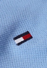 Světle modrá pletená tkanina s malým obdélníkovým vyšitým logem obsahujícím černé, bílé a červené horizontální barevné bloky.