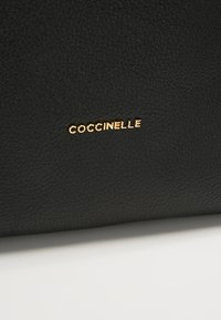 Musta nahkalaukku, jossa on teksturoitu pinta; esillä on suurikokoinen kultainen logo, jossa lukee "COCCINELLE". Minimalistinen muotoilu ilman ylimääräisiä yksityiskohtia.