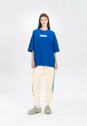 Joven mujer de pie contra un fondo blanco, lleva una camiseta azul de gran tamaño, pantalones deportivos crema con estampados laterales y zapatillas beige con textura.