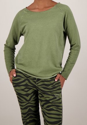 Person trägt olivgrünes Langarmshirt und grün-schwarze Zebramuster-Hose, Hände in den Taschen, vor einfarbigem Hintergrund.