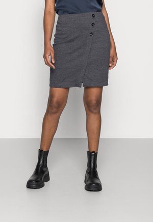 Femme debout portant une mini-jupe boutonnée gris foncé, un haut noir et des bottines noires sur un sol gris clair avec un fond blanc.