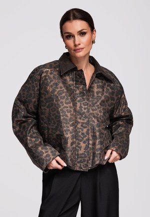 Žena má na sobě koženou bundu s hnědočerným leopardím vzorem a širokým límcem, černé kalhoty, stojí před jednobarevným světlým pozadím.