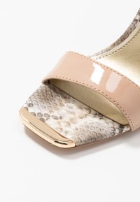 Beige högklackad sandal med en glansig lackrem, ormskinnsstruktur på sulan och en guldfinish på tån.