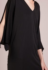 Svart klänning med V-ringning, korta breda ärmar med öppna sidor, flytande textur och en smidig, figurnära silhuett.