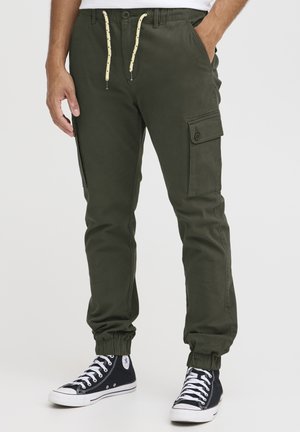 Olivegrønne cargo-bukser med elastiske manchetter, to sidelommer og en frontlår med gul snor. Matchende med sorte sneakers.