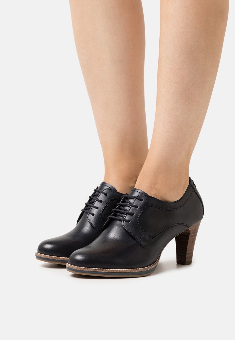 Zapatos de cuero negro con tacón, puntera redonda, diseño con cordones y parte superior texturizada. Cuentan con un tacón de madera apilada y costuras en contraste.