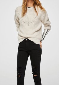 Suéter crema con un patrón de punto texturizado y un diseño en forma de V, encima de una camisa de manga larga a rayas en blanco y negro, combinado con jeans negros rasgados.