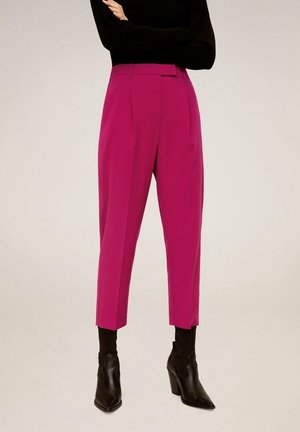 Femme portant un pantalon court taille haute rose vif associé à des bottines noires à talons et un haut noir à manches longues, bras croisés.