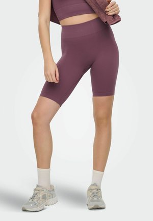 Woman wearing high-waisted, ribbed purple bike shorts, matching crop top, white socks, and gray sneakers, holding a folded matching fabric.