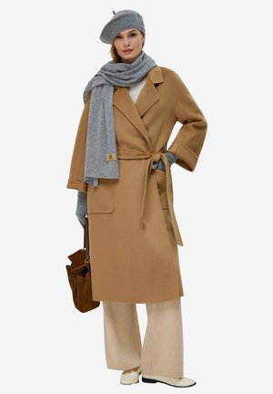 Frau trägt camel-farbenen Mantel mit Gürtel, graue Baskenmütze, Schal, Handschuhe, cremefarbene Hosen mit weitem Bein, weiße Schuhe, hält eine braune Handtasche, vor einfarbigem Hintergrund.