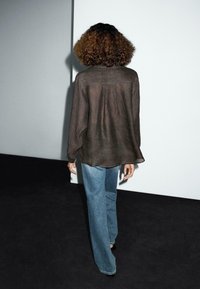 Chemise longue en voile marron à manches longues avec une coupe décontractée et un pli au dos, associée à un jean large bleu clair sur un fond sombre.