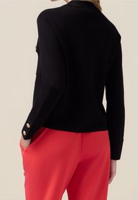 Svart kavaj med skräddarsydd passform, som har en notchad krage och guldknappar på ärmarna, tillsammans med röda byxor.