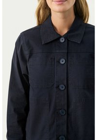 Ciemnoniebieska kurtka zapinana na guziki, wykonana z bawełnianej tkaniny, z kołnierzem, dwiema kieszeniami na piersi oraz zapięciem na cztery okrągłe guziki.