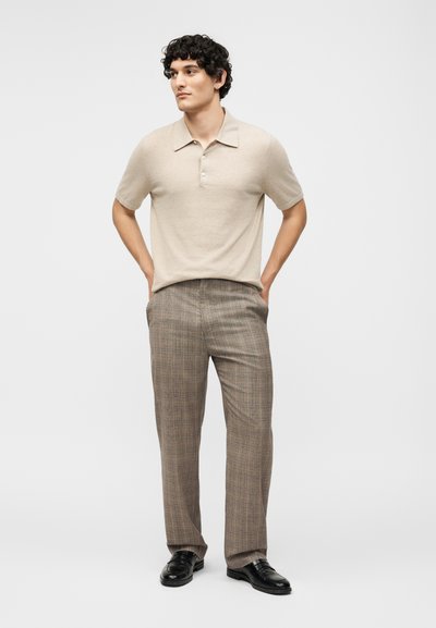 Νέος άνδρας με σγουρά σκουρόχρωμα μαλλιά, φοράει μπεζ πόλο με κοντά μανίκια, καφέ καρό παντελόνια, μαύρα παπούτσια, στέκεται με τα χέρια στις τσέπες μπροστά σε λευκό φόντο.